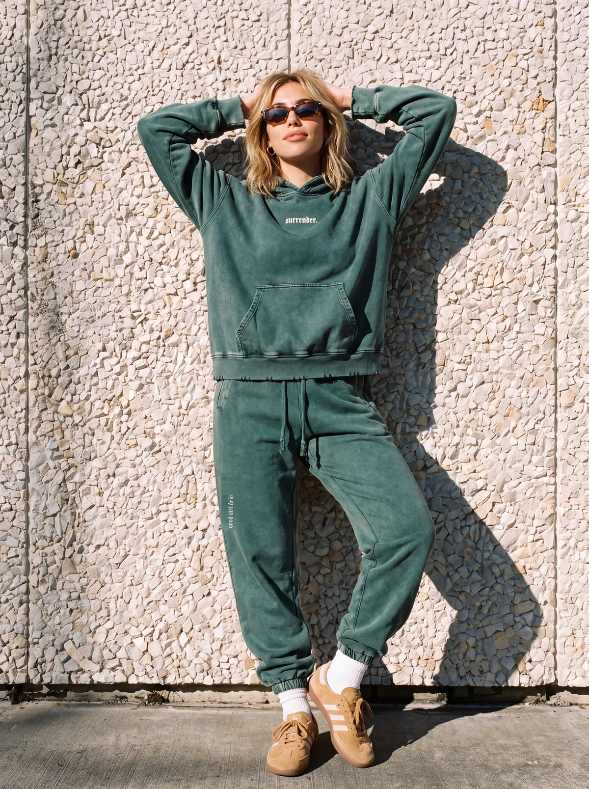 Woman in the green "surrender." snow wash hoodie and matching joggers set against a pebble wall