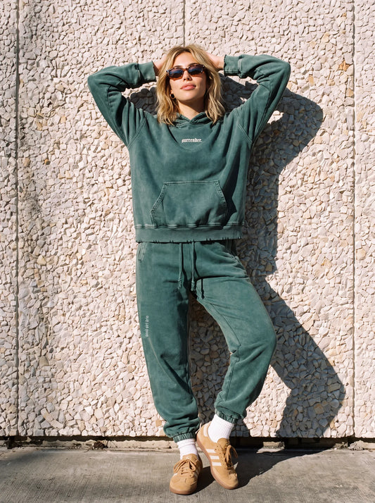 Woman in the green "surrender." snow wash hoodie and matching joggers set against a pebble wall